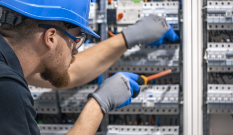 A male electrician works in a switchboard with an electrical connecting cable. Electrician with screwdriver tightens electrically operated switching equipment in fuse box.